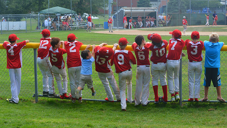 A group of boys from Milton National baseball standing on a fence watching a baseball game, excited for their upcoming Cooperstown trip.