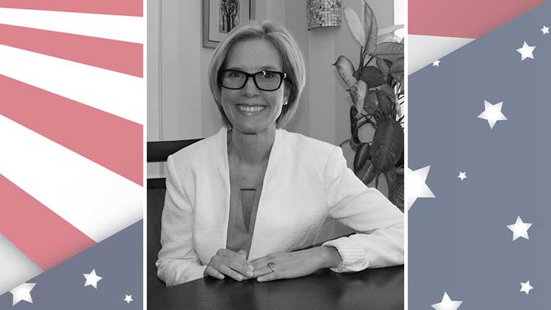 A woman in glasses is sitting at a table in front of an american flag.