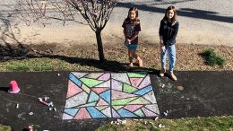 Two girls from Milton sharing their spring artwork as they stand next to a chalk drawing on the sidewalk.