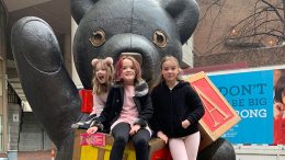 Three girls posing in front of a teddy bear statue.