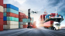 A truck is driving through a container yard with a plane in the background, highlighting the impact of supply chain issues on small businesses importing materials.