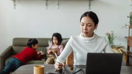 A woman is writing on a laptop while sitting at a table with her children, balancing work and family schedules during the summer.