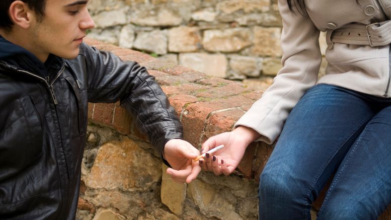 A teen offers a cigarette to a woman perched upon a wall.