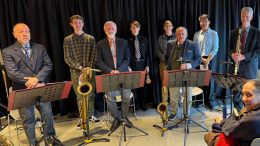A group of male musicians with various saxophones standing and sitting by their music stands at Milton Public Library, possibly preparing for a performance or rehearsal as part of the library's adult programming for April
