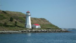 A traditional white lighthouse with a red top on a rocky shoreline with a small Milton Public Library nearby, under a clear blue sky.
