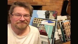 Man smiling with a collection of colorful artwork from the "April Art Extravaganza: Don Hammontree's Vibrant Exhibit at Wotiz Gallery" leaning against a brick wall.