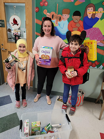 Two children and an adult standing in a hallway with donated items for Ramadan in Milton 2024, smiling at the camera.