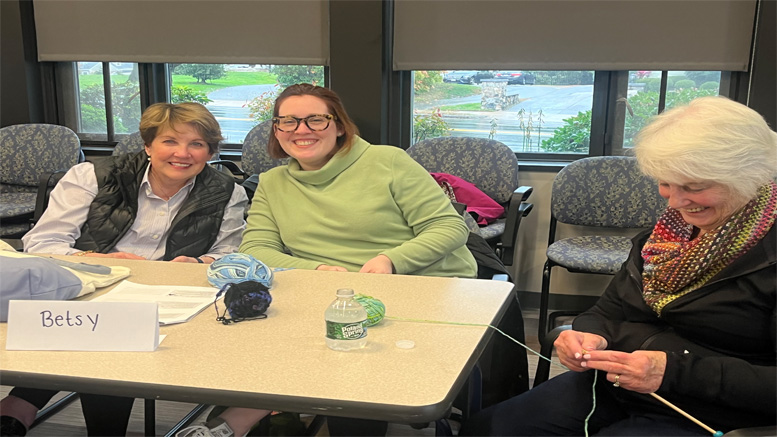 Three women smiling around a table with papers and water bottles, one knitting, inside a well-lit room with windows for a special screening of 'The Holdovers' on May 15.