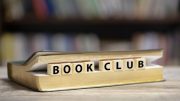 A book lies open with letter tiles spelling "Celebrity Book Club" laid across its pages, set against a blurred bookshelf background.