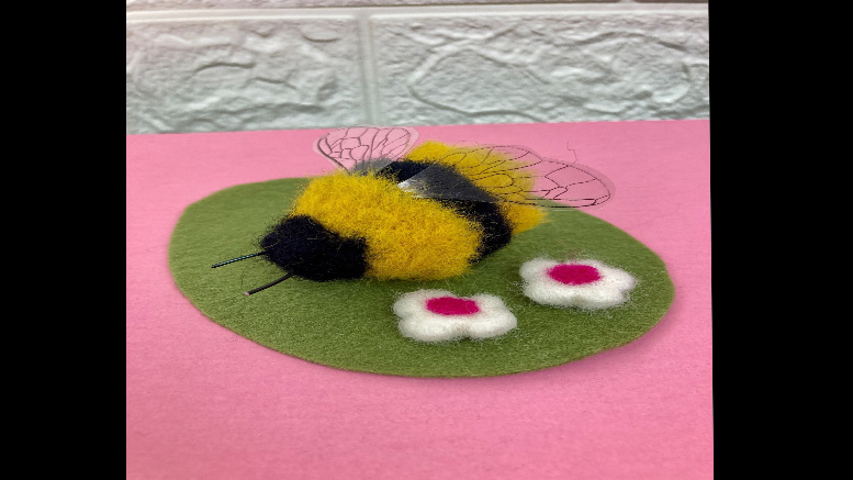 A crafted felt bee on a green oval mat with two white and pink flowers, set against a pink background with a white tiled pattern, invites you to discover Chair Yoga with Marianne Zullas on