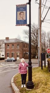 A woman in a pink jacket stands smiling beneath a banner of a U.S. Army soldier attached to a lamp post on a quiet suburban street.