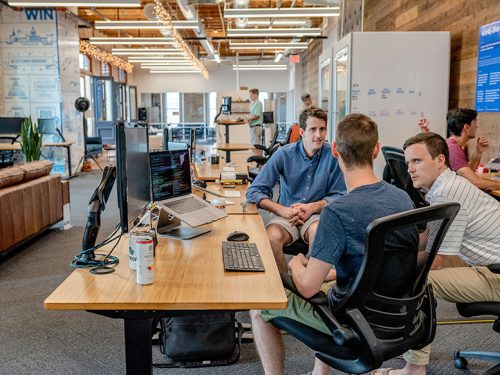 Colleagues discussing the European job market at a wooden table in a busy open-plan office with multiple computer screens and whiteboards.