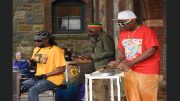 Three musicians wearing colorful clothing are performing outdoors. One plays a drum set, another plays congas, and the third plays a steelpan. A brick and stone building stands in the background. Experience holiday magic at Eustis Estate on December 14: an evening of festive cheer awaits you there!
