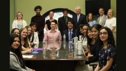 A group of people sits and stands around a conference room table, posing for a photo. Some are holding papers, while others look at the camera. The room has a large window with a decorative circular design—a backdrop that underscores their commitment to initiatives like Fresh Truck Mobile Market in Boston.