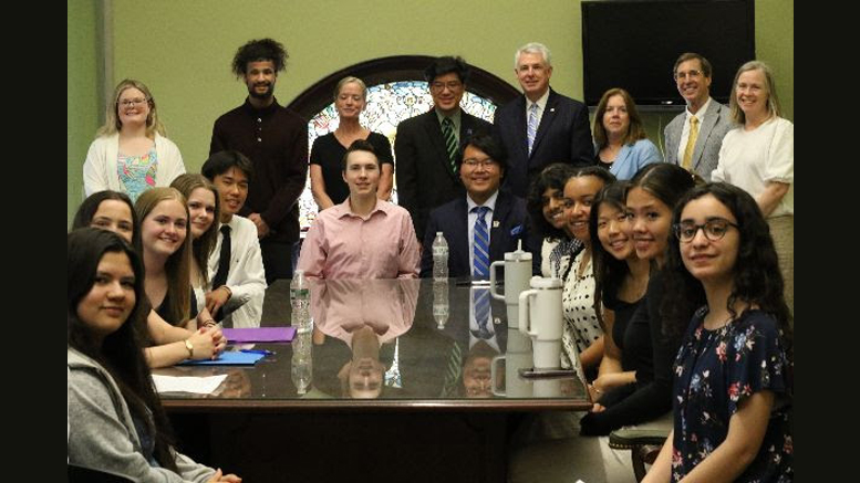 A group of people sits and stands around a conference room table, posing for a photo. Some are holding papers, while others look at the camera. The room has a large window with a decorative circular design—a backdrop that underscores their commitment to initiatives like Fresh Truck Mobile Market in Boston.