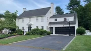 A two-story white house with black shutters and a black garage door. Two workers from Costa Painting Services are transforming your home as they paint the exterior. The front yard has green grass and bushes, with a ladder visible on the left side.
