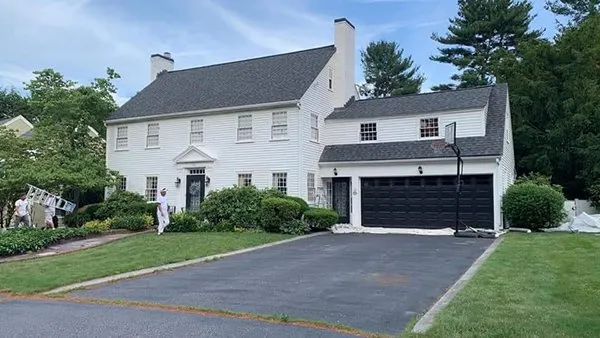A two-story white house with black shutters and a black garage door. Two workers from Costa Painting Services are transforming your home as they paint the exterior. The front yard has green grass and bushes, with a ladder visible on the left side.