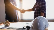 Two people shake hands in an office setting with construction plans, a white hard hat, and a small house model on a table.
