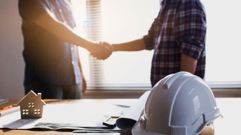 Two people shake hands in an office setting with construction plans, a white hard hat, and a small house model on a table.