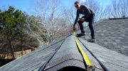Person measures roof ridge with yellow tape measure under clear sky.