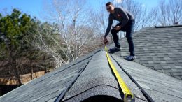 Person measures roof ridge with yellow tape measure under clear sky.