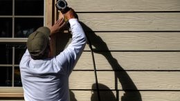 A man wearing a cap uses a power tool on the siding of a house.