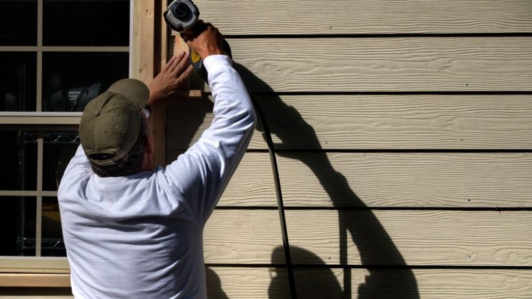 A man wearing a cap uses a power tool on the siding of a house.