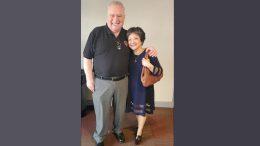 A man and a woman stand indoors, both smiling warmly. The man sports a black polo with gray pants while the woman wears a dark dress, clutching a brown handbag. Perhaps they're discussing the exciting adult programming Milton Public Library announces for October 2023.