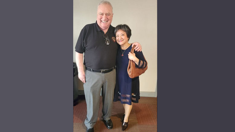 A man and a woman stand indoors, both smiling warmly. The man sports a black polo with gray pants while the woman wears a dark dress, clutching a brown handbag. Perhaps they're discussing the exciting adult programming Milton Public Library announces for October 2023.
