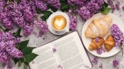 Open book, cappuccino with heart latte art, and croissants on a plate surrounded by lilac flowers.