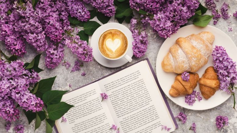 Open book, cappuccino with heart latte art, and croissants on a plate surrounded by lilac flowers.