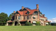 Experience the charm of a large stone and brick mansion with red roofs, chimneys, and striped awnings set against a clear blue sky. Join us on Mar. 23 for the Wintersounds Series at Milton's Eustis Estate amid the lush grassy lawn.