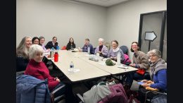 In a meeting room, a group of twelve people sits around a conference table, smiling amidst their personal belongings. They're busy planning as the Milton Public Library announces its exciting adult programs for April 2025.
