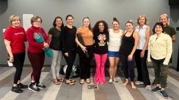 A group of eleven people standing indoors, casually dressed, some holding yoga mats, smiling at the camera, embodying the community spirit as they celebrate The Milton Public Library's announcement of adult programs for April 2025.