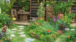 stunning gardens in a lush scene featuring a stone path, vibrant flowers, and greenery. A wooden table and chairs sit cozily in the corner, while a wooden fence entwined with climbing plants