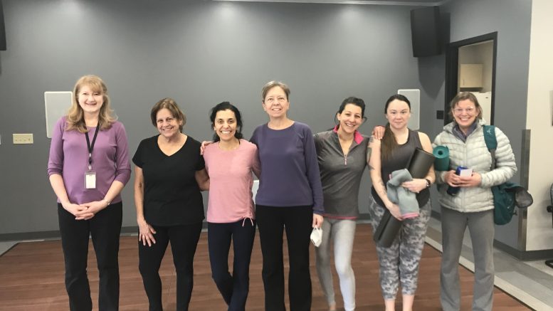 Seven women stand in a yoga studio, some holding yoga mats and water bottles, all smiling at the camera. The room has wooden floors and gray walls—part of the Milton Public Library’s adult programs in May 2025.
