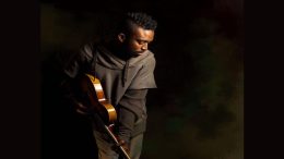 A person stands holding an electric guitar, looking down against a dark, shadowy background—an introspective mood that echoes the creative solitude found at a writing retreat, like those held at Milton's Eustis Estate in April 2025.