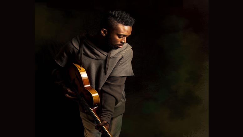 A person stands holding an electric guitar, looking down against a dark, shadowy background—an introspective mood that echoes the creative solitude found at a writing retreat, like those held at Milton's Eustis Estate in April 2025.