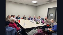 A group of twelve people sit around a rectangular table in a meeting room at Milton Public Library, some knitting and others smiling at the camera during the Knit & Chat—one of May events you don’t want to miss.