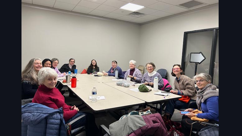 A group of twelve people sit around a rectangular table in a meeting room at Milton Public Library, some knitting and others smiling at the camera during the Knit & Chat—one of May events you don’t want to miss.