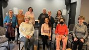 A group of ten adults, mostly older women, pose for a photo in a gray-walled room after a Chair Yoga Stretch & Strengthen class with Marianne Zullas; two people are seated upfront while others stand behind.
