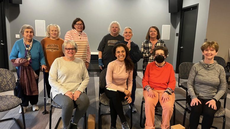 A group of ten adults, mostly older women, pose for a photo in a gray-walled room after a Chair Yoga Stretch & Strengthen class with Marianne Zullas; two people are seated upfront while others stand behind.