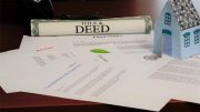 A rolled-up title and deed document, final notice letter, assorted papers, and a small model house are placed on a desk—reminding homeowners about the risk of deed fraud and the benefits of a free alert service.