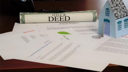 A rolled-up title and deed document, final notice letter, assorted papers, and a small model house are placed on a desk—reminding homeowners about the risk of deed fraud and the benefits of a free alert service.