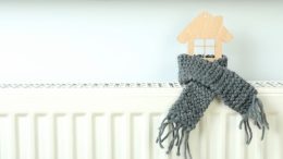 A wooden house shape, wrapped in a knitted gray scarf on a white radiator, evokes warmth and energy efficiency. Mass residents, don't miss winter heating assistance—apply by April 30 to ensure cozy comfort all season long.