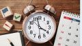 A bronze twin-bell alarm clock shows nearly 12:00, serving as a reminder to work smarter, not harder. Nearby, a calendar, notepad, and wooden blocks with icons like flags and checkmarks create an organized Small Business Corner on the wooden surface.