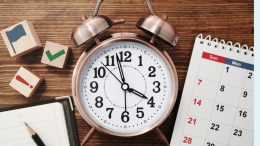 A bronze twin-bell alarm clock shows nearly 12:00, serving as a reminder to work smarter, not harder. Nearby, a calendar, notepad, and wooden blocks with icons like flags and checkmarks create an organized Small Business Corner on the wooden surface.