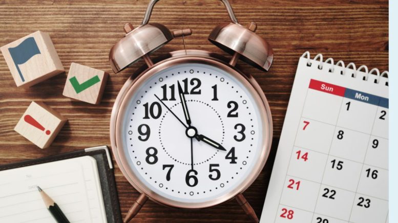 A bronze twin-bell alarm clock shows nearly 12:00, serving as a reminder to work smarter, not harder. Nearby, a calendar, notepad, and wooden blocks with icons like flags and checkmarks create an organized Small Business Corner on the wooden surface.