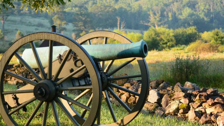 A historic cannon stands on a grassy field near a pile of rocks, with trees under a clear sky setting the stage for an experience of "Glory" on the big screen this April 23, 2025.