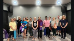 A group of people of various ages stand in a room at the Milton Public Library, holding yoga mats and posing for a group photo before or after a Free Yoga class this Summer.